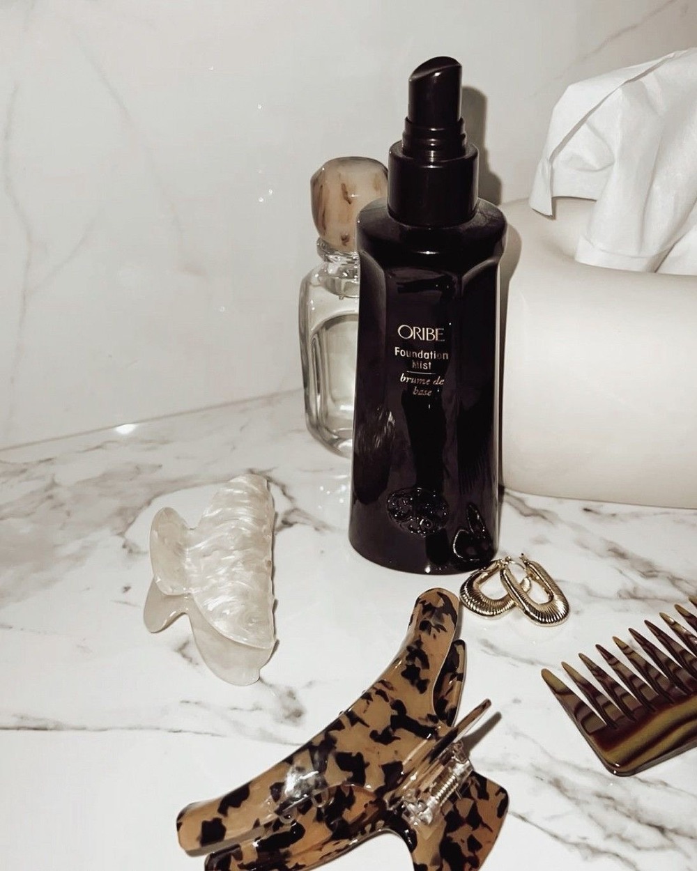 Oribe Foundation Mist with tortoise clips and gold earrings on marble